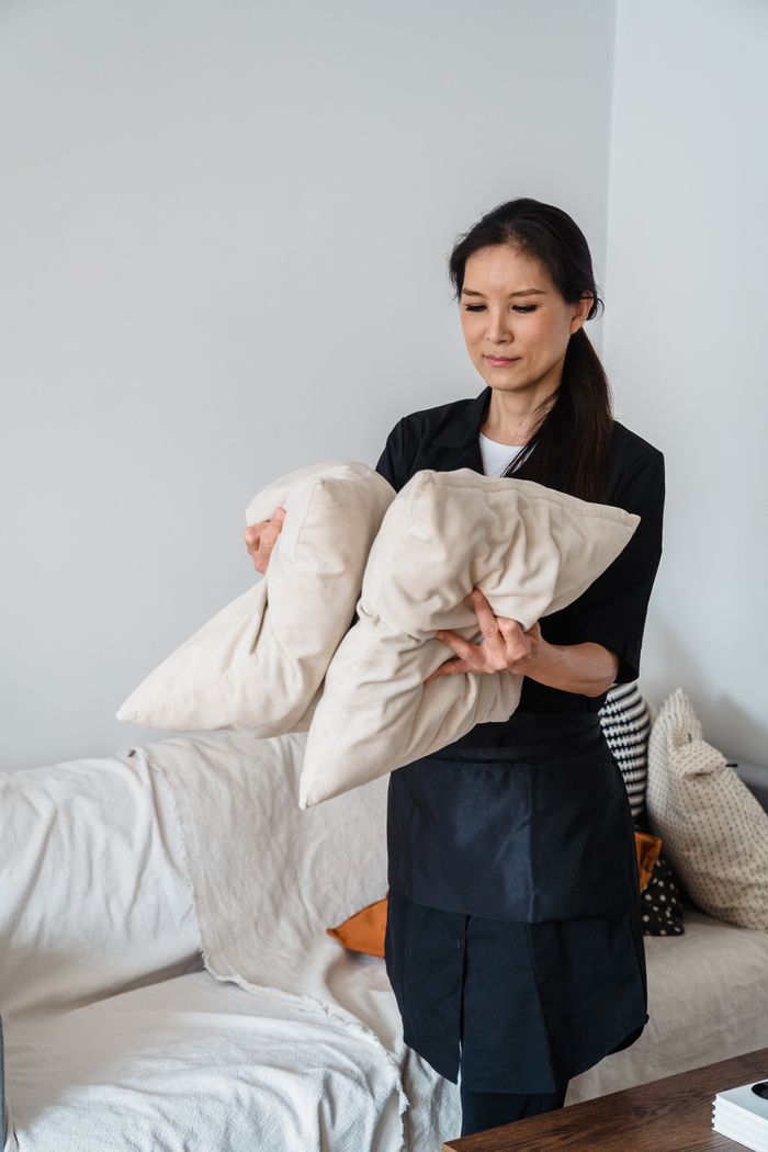 A cheerful housekeeper tidying up cushions on a sofa in a sunlit living area.